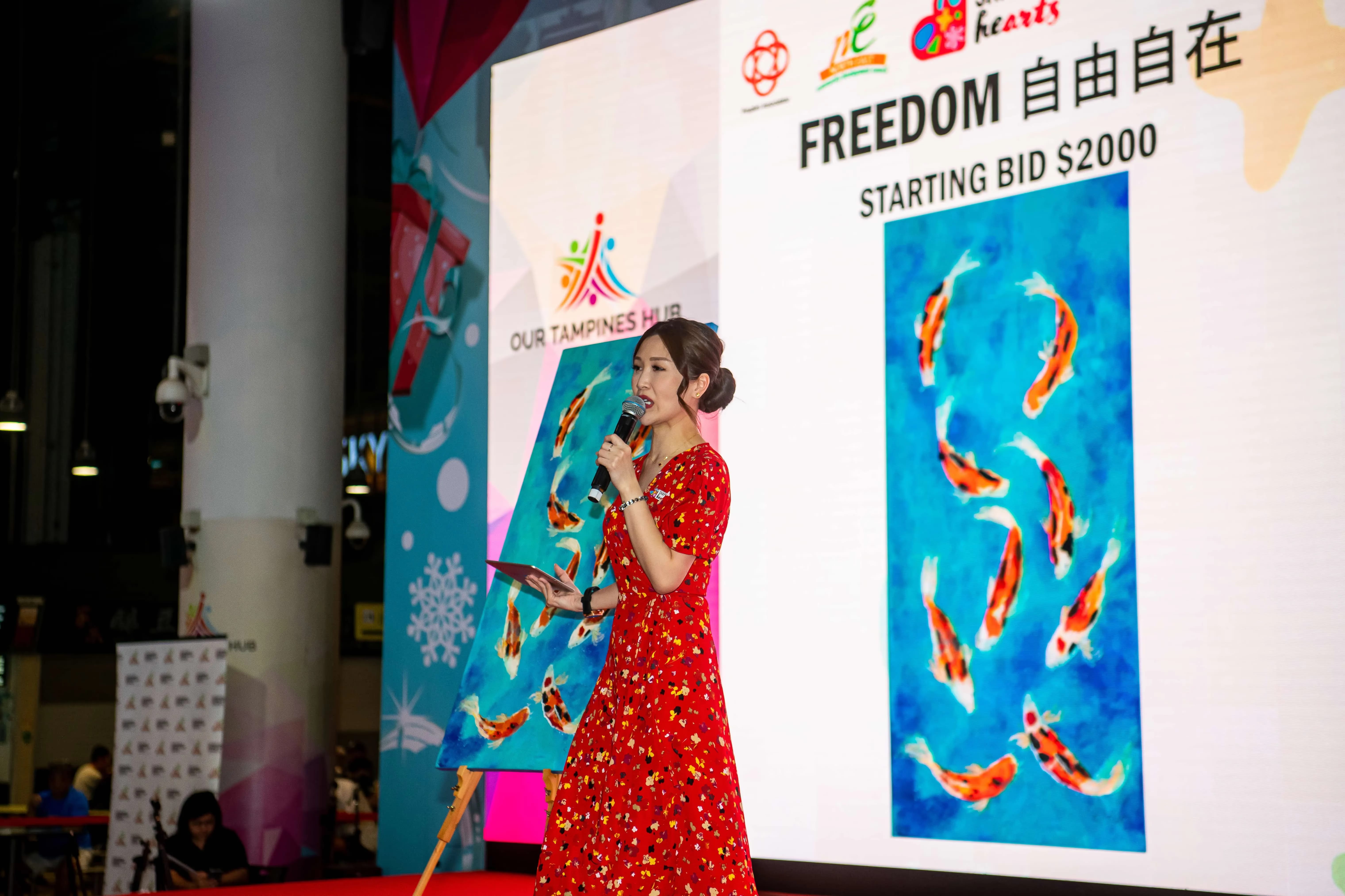 A person in a red dress is presenting a painting of koi fish with a starting bid of $2000 at Our Tampines Hub.
